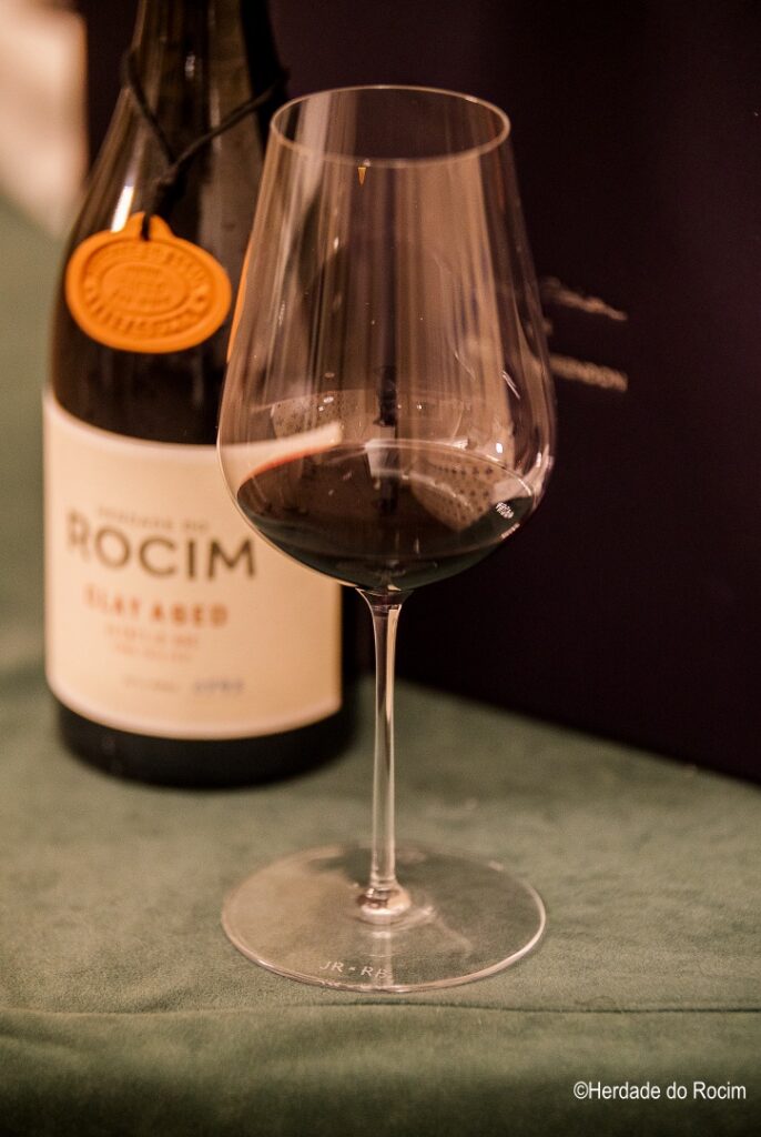 Foto aproximada de copo de vinho criado por Jancis Robinson, com vinho tinto no fundo e garrafa de vinho ao lado.